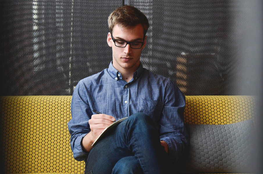 A young man, in his early 20s, sat on a yellow seat writing in a notebook. The man is wearing black glasses, a denim shirt, and blue jeans.